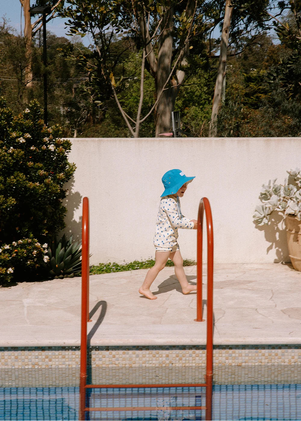 A child wearing a white patterned swimsuit and a bright blue sun hat walks barefoot along a poolside deck next to rust-colored ladder rails, with a white wall and trees in the background.