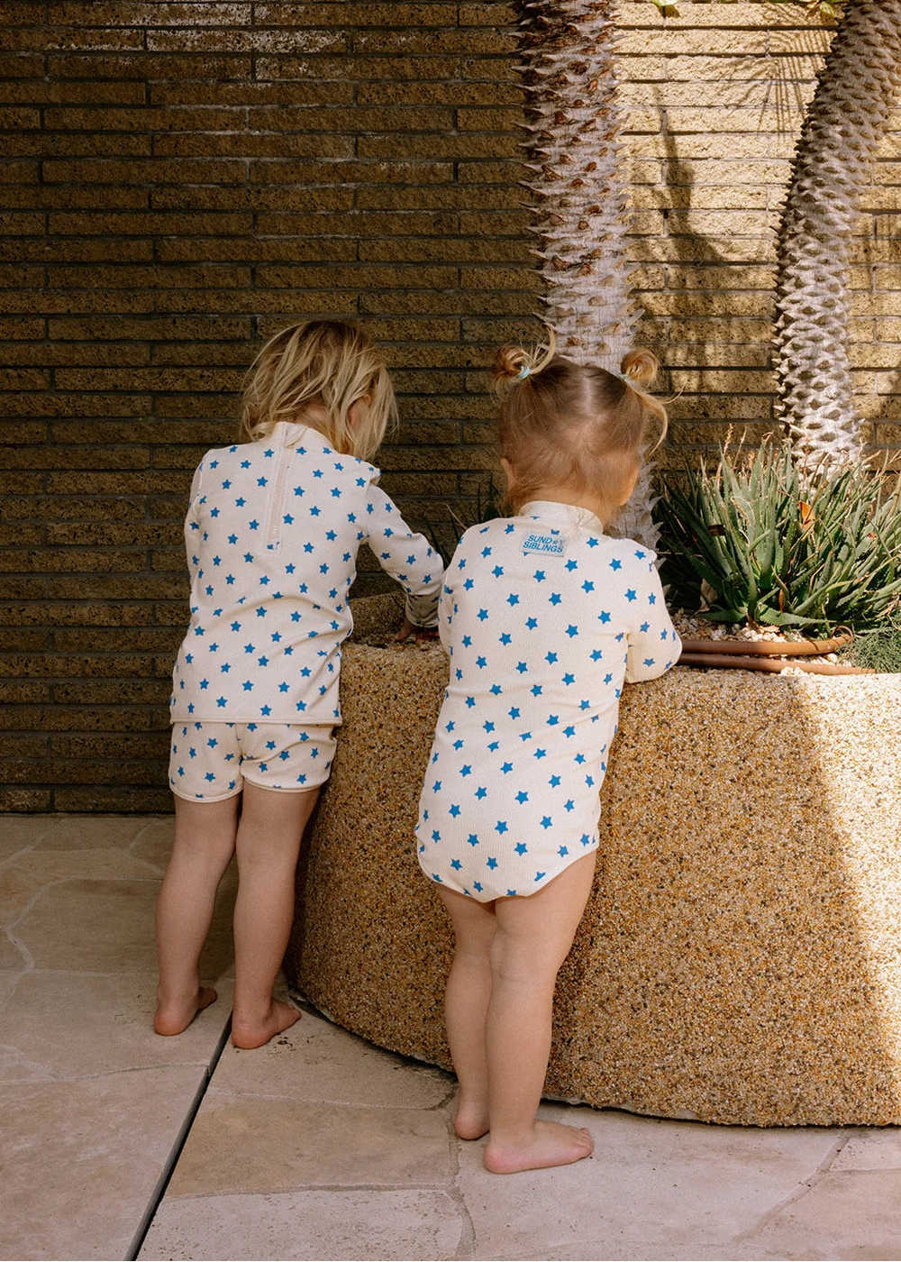 Two young children wearing matching white long-sleeve bodysuits with blue star patterns, standing barefoot outdoors next to a textured stone planter against a brick wall.