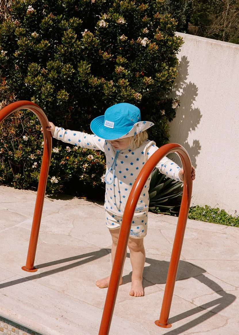 A toddler stands barefoot on a pool deck wearing a white one-piece swimsuit with blue polka dots and a bright blue sun hat, holding onto orange pool ladder rails in front of a large green bush and white wall.
