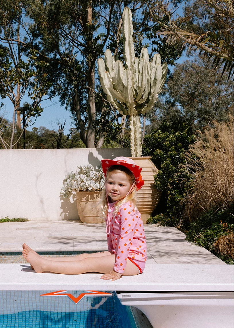 A young child wearing a pink and red polka-dot swimsuit and a red sun hat sits on the edge of a swimming pool outdoors, with trees, a cactus, and a white wall in the background.