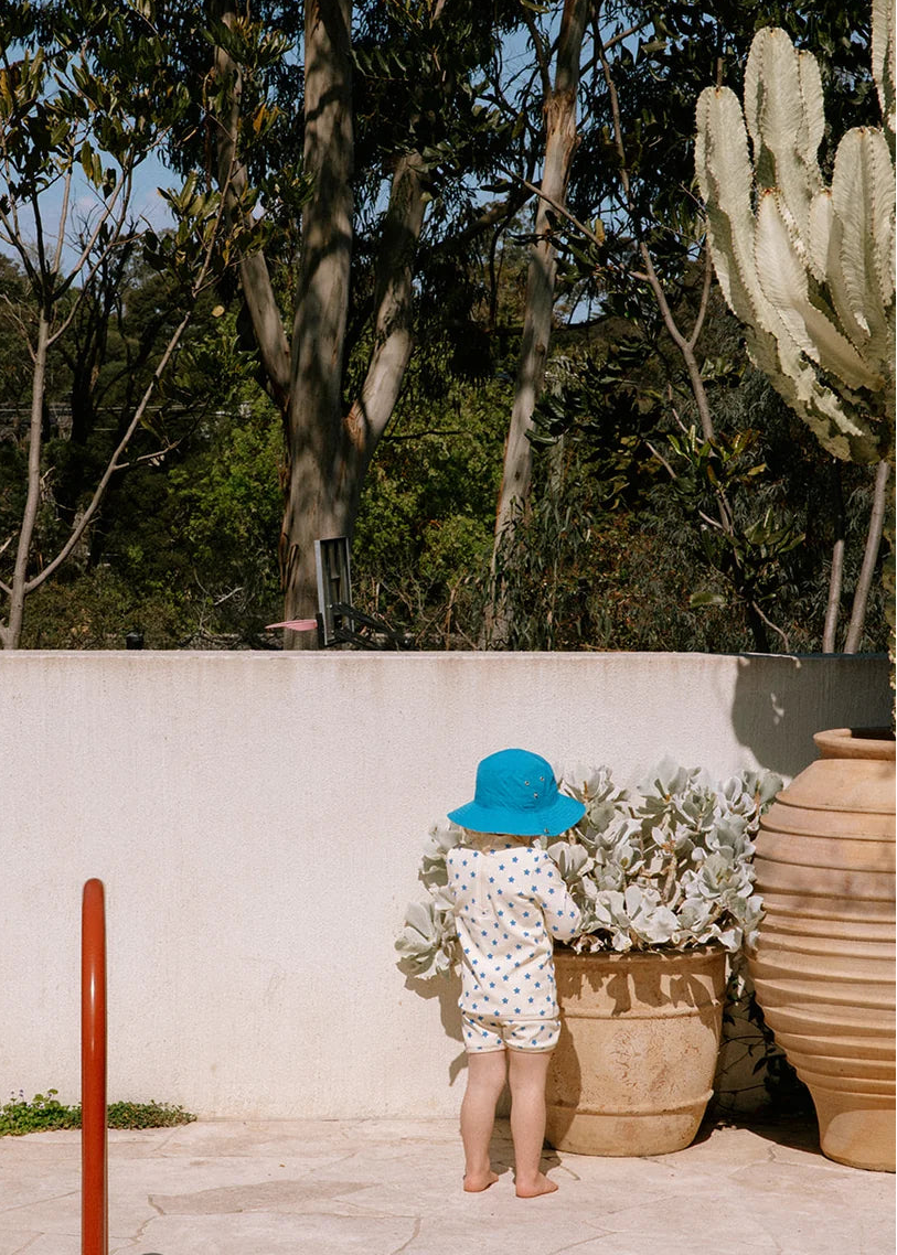 A toddler stands barefoot outdoors near a beige wall, wearing a white patterned romper and a bright blue sun hat, with potted plants and tall trees in the background.