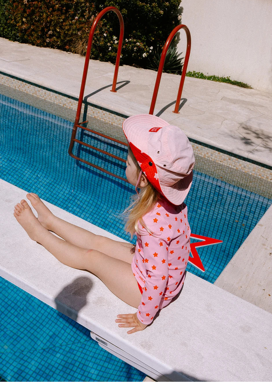 A young child sitting on the edge of a swimming pool, wearing a pink sun hat with red accents and a pink long-sleeve swimsuit covered in small red stars.