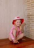 A young girl with blonde hair sits barefoot on a wooden floor in the corner of a room, wearing a pink long-sleeve swimsuit with red star patterns and a matching pink sun hat with a red brim.