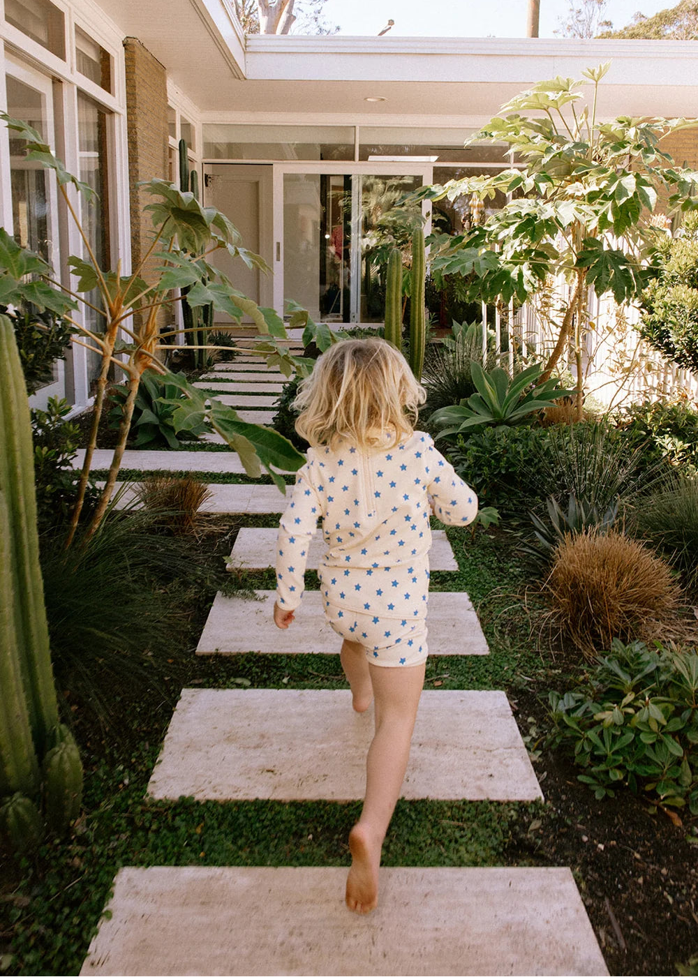 A young child with blonde hair is seen from behind, running barefoot down a stone path toward a house, wearing a cream long-sleeve romper covered in small blue and yellow polka dots.
