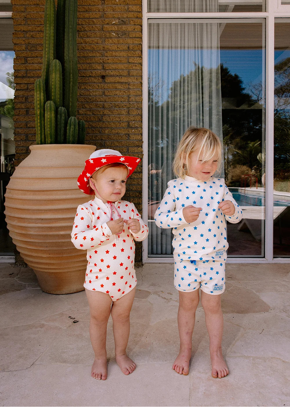 Two toddlers standing outdoors on a patio in matching red and blue polka dot swim sets, with one wearing a red polka dot sun hat. A large terracotta planter with cacti and glass patio doors are visible in the background.