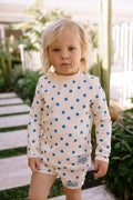 A toddler with blonde hair stands outdoors wearing a cream long-sleeve romper covered in blue star patterns. The brand name 'JONATHAN' is visible on a small patch near the leg.