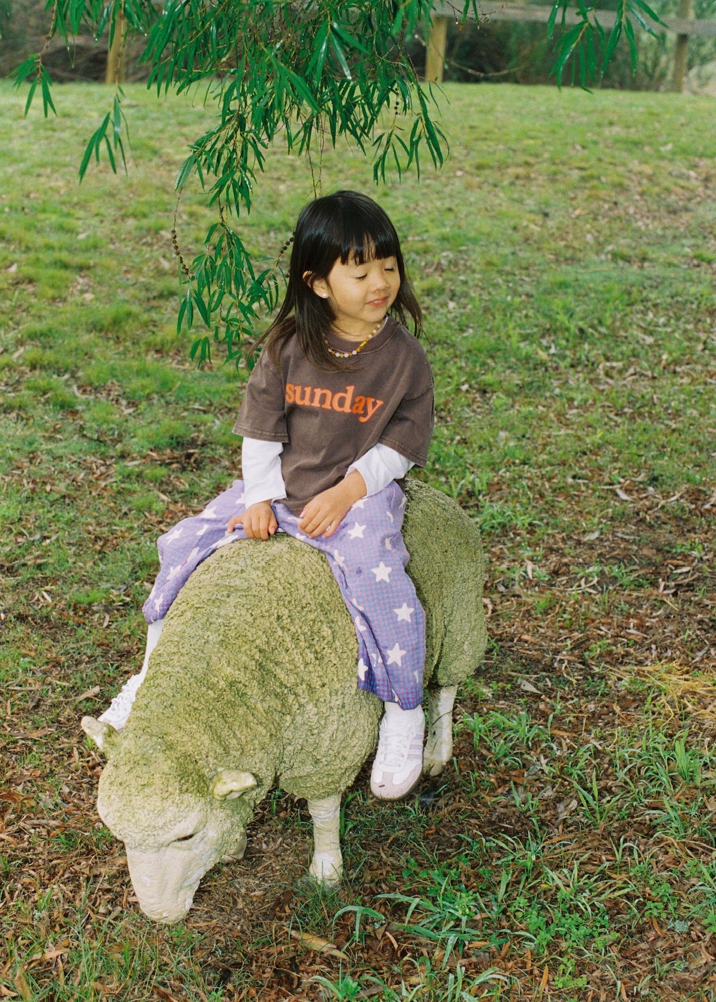 A young girl with dark hair sits on a sheep-shaped garden statue in a grassy outdoor area. She is wearing a brown long-sleeve shirt with orange text that reads 'sunday' and a purple skirt with white stars.