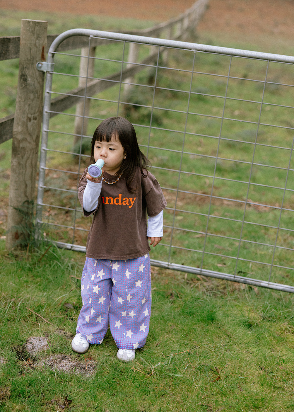 A young child wearing a brown t-shirt with orange text and purple pants with white stars stands in a grassy field next to a metal gate.