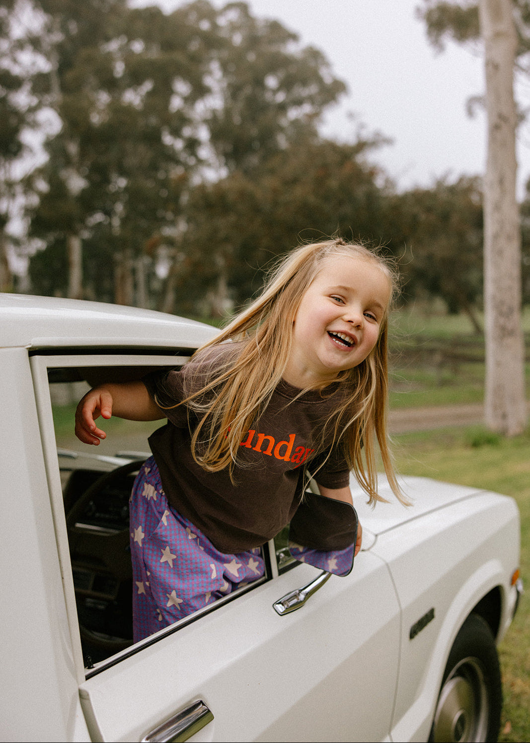 A young girl with long blonde hair leans out of the window of a white vintage car, smiling and laughing. She is wearing a dark brown t-shirt with orange text and purple star-patterned pants.