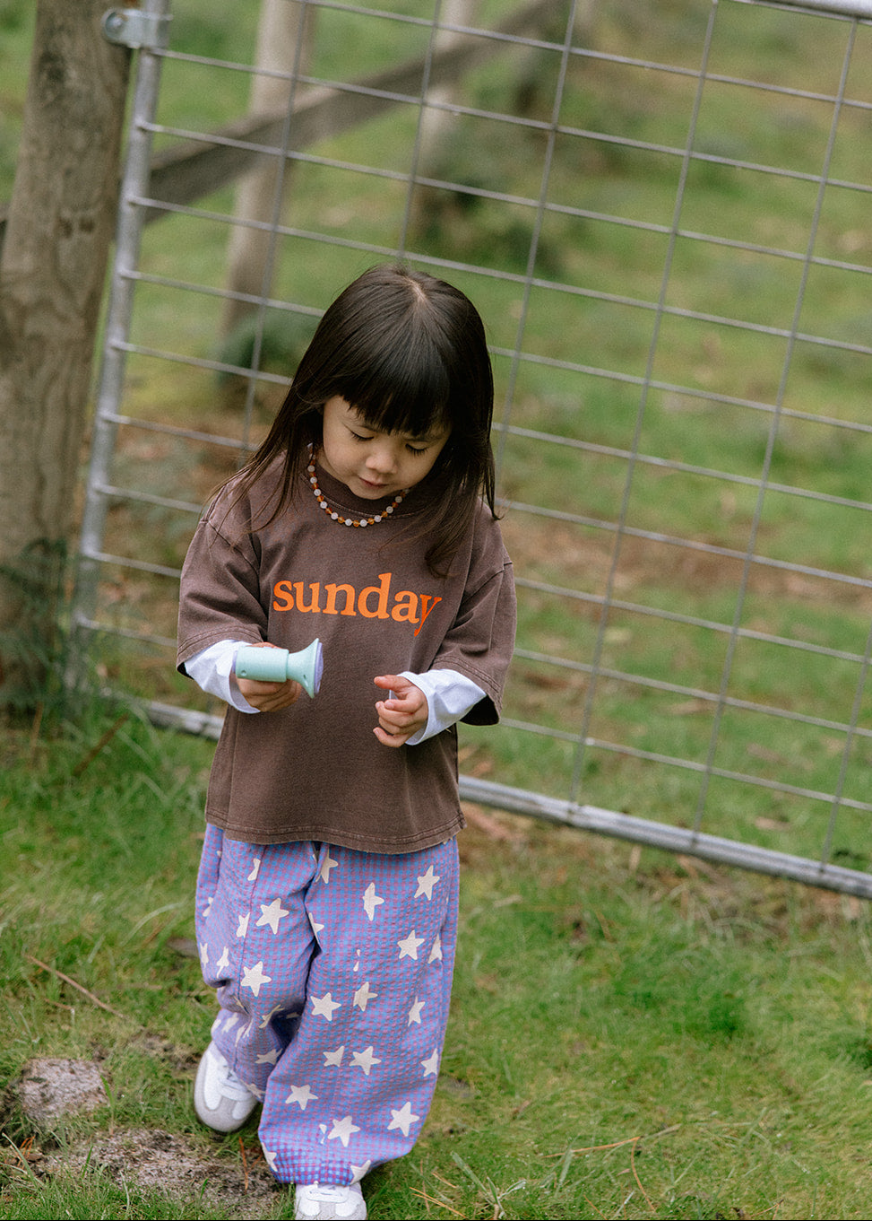 A young girl with dark hair stands outdoors in front of a wire fence, wearing a brown t-shirt with orange 'sunday' text and purple star-patterned pants.