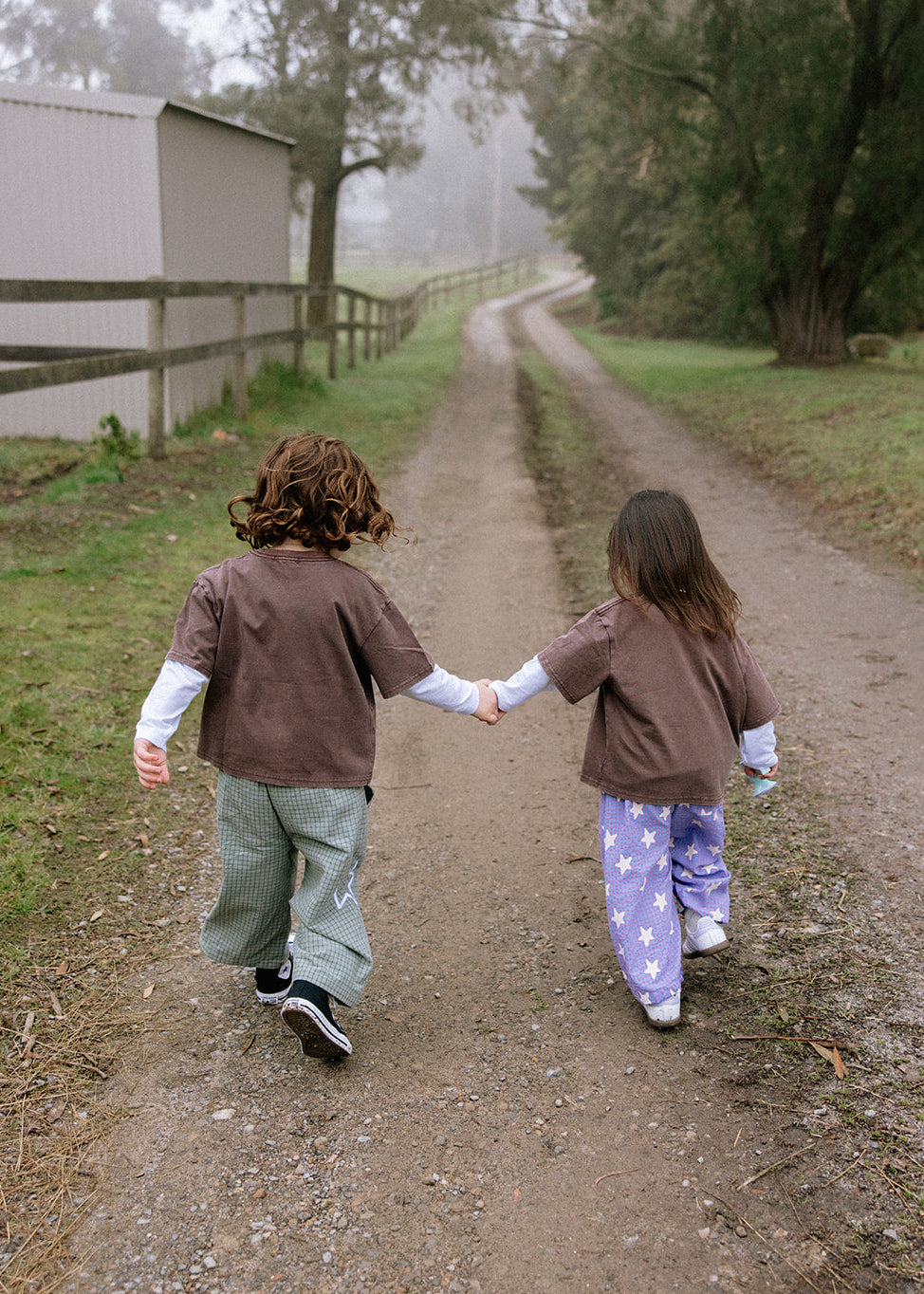 Two children holding hands walking away on a muddy dirt path, wearing matching brown t-shirts, one in green pants and the other in purple starry pajama pants, with a farm scene in the background.