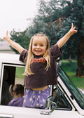 A young girl with blonde hair stands in the open window of a white vehicle, smiling with her arms raised. She is wearing a dark brown t-shirt with orange text and purple patterned shorts.