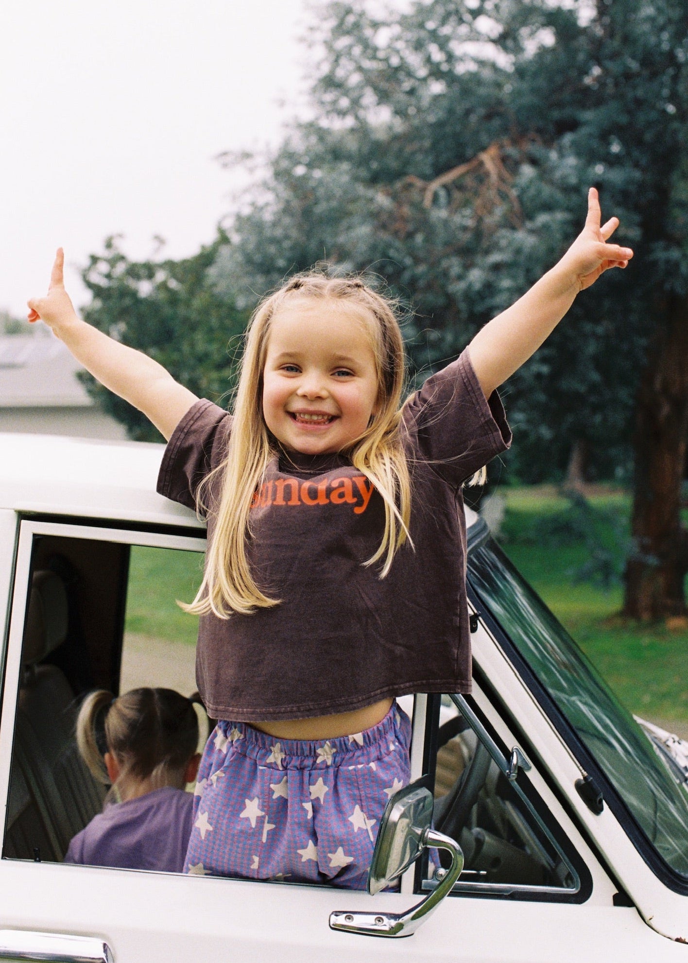 A young girl with blonde hair stands in the open window of a white vehicle, smiling with her arms raised. She is wearing a dark brown t-shirt with orange text and purple patterned shorts.