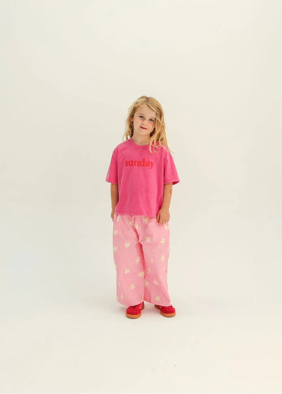 A young girl stands against a plain white studio background wearing a bright pink short-sleeve t-shirt with red text and light pink patterned pants with red shoes.