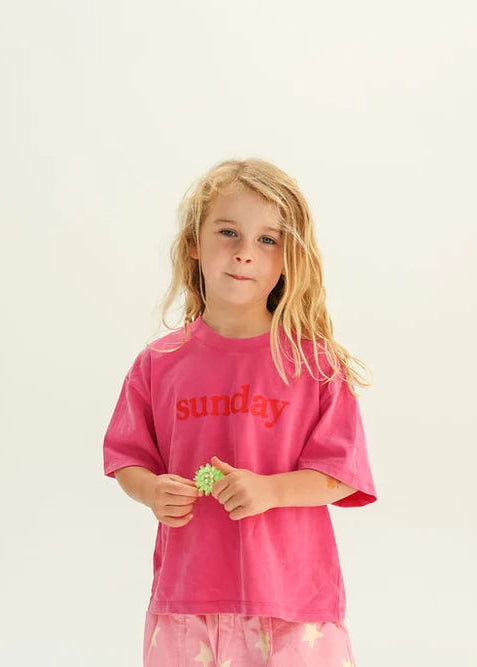 A young girl with blonde hair stands against a plain white studio background wearing a bright pink t-shirt with the word 'Sunday' printed in red across the chest.