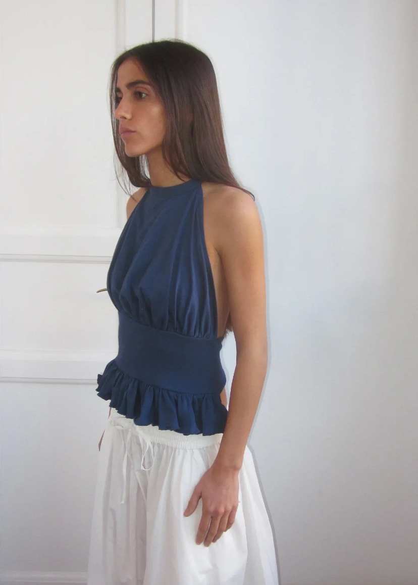 Woman wearing a blue top with ruffled details and white pants against a plain background