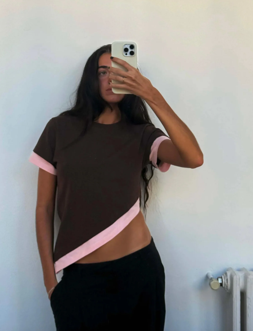 Person wearing a brown and pink top taking a mirror selfie against a white wall.