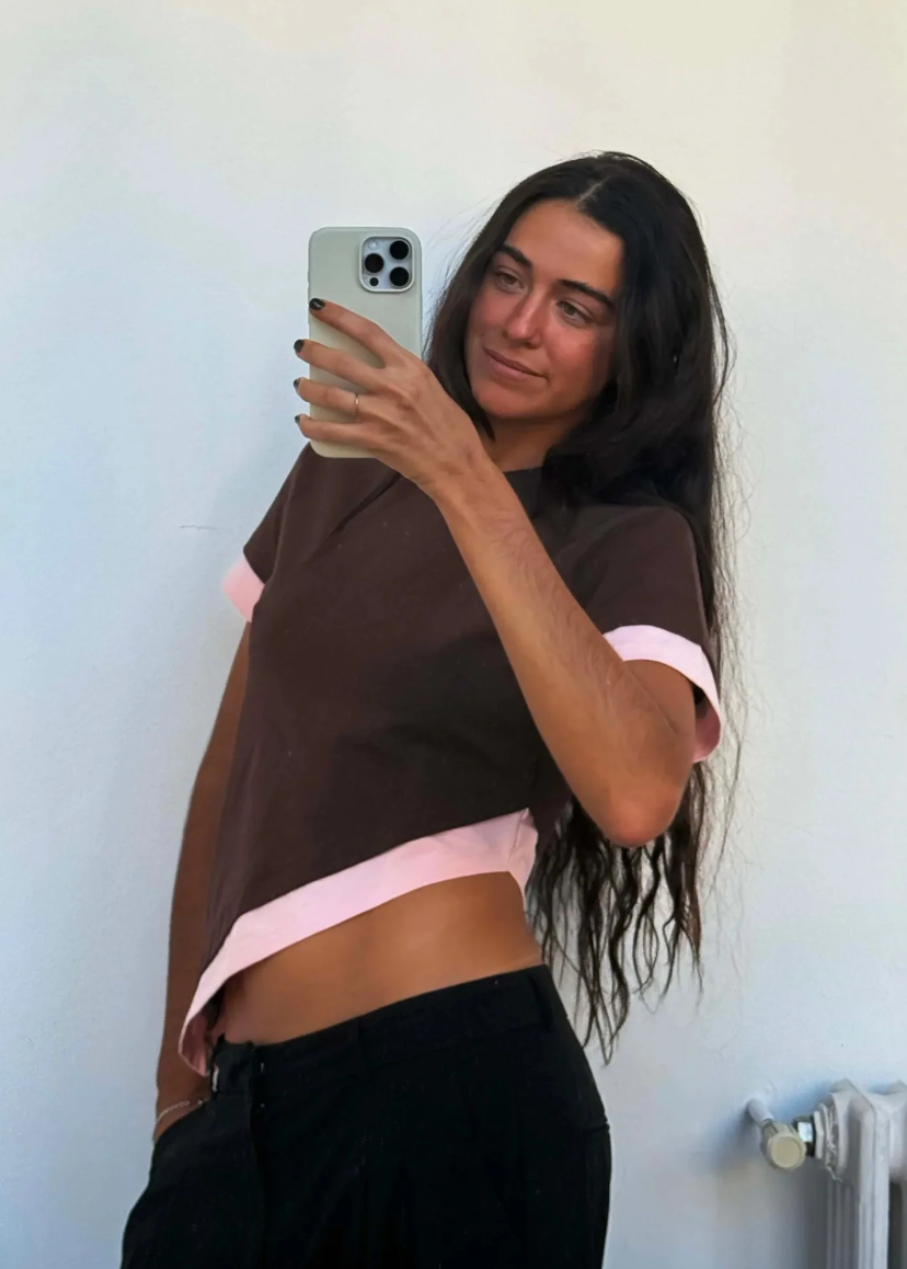 Person taking a mirror selfie wearing a brown and pink top against a light blue wall.