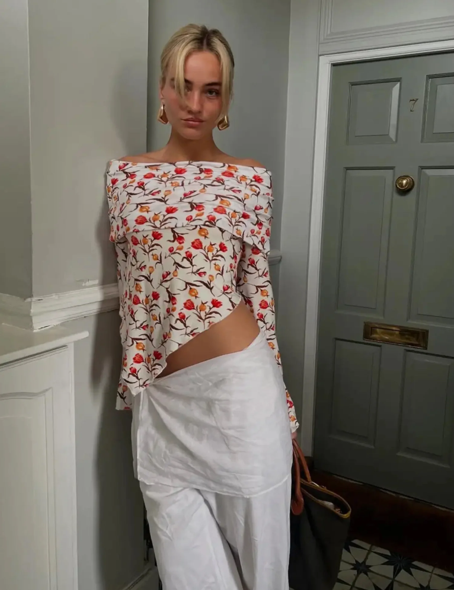 Woman wearing a floral off-shoulder top and white pants in a room with a door.