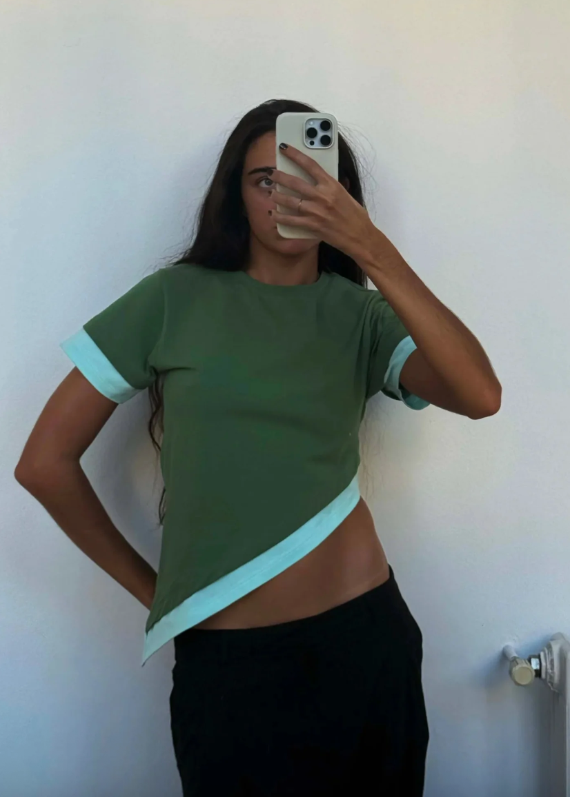 Person wearing a green and blue top taking a mirror selfie against a white wall.
