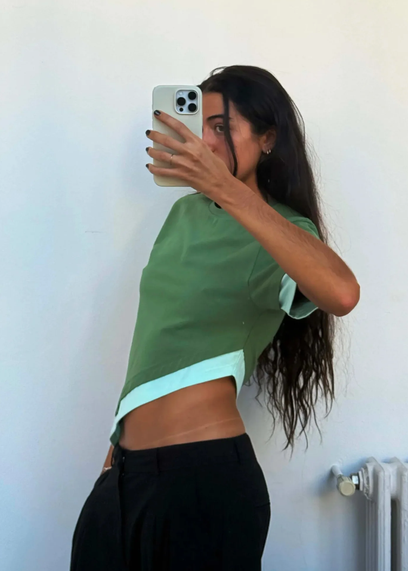 Person taking a mirror selfie wearing a green crop top and black pants against a white wall.