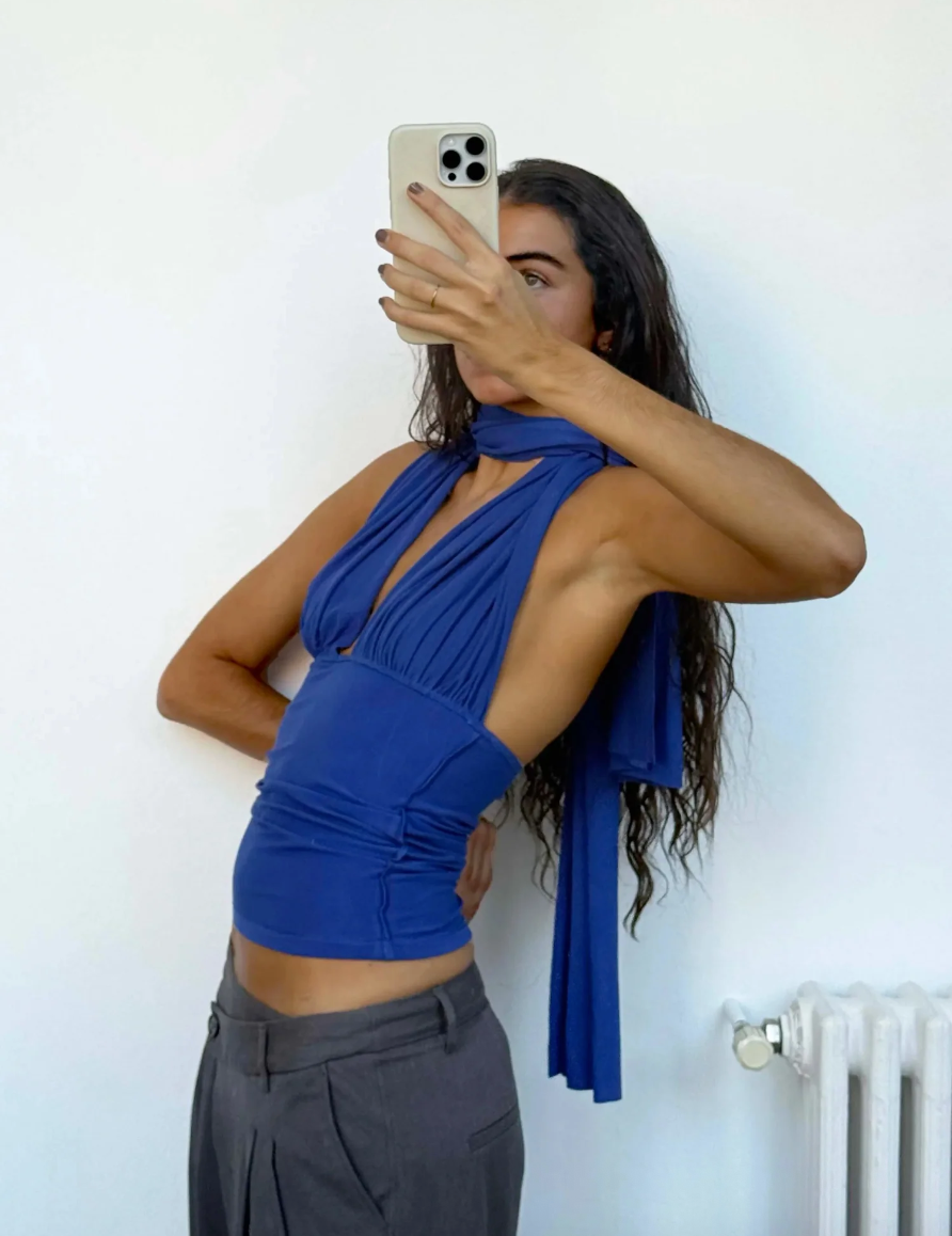 Woman taking a mirror selfie wearing a blue top and gray pants against a white wall.