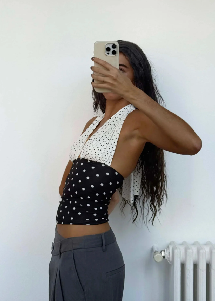 Person wearing a black and white polka dot top with gray pants against a white wall.