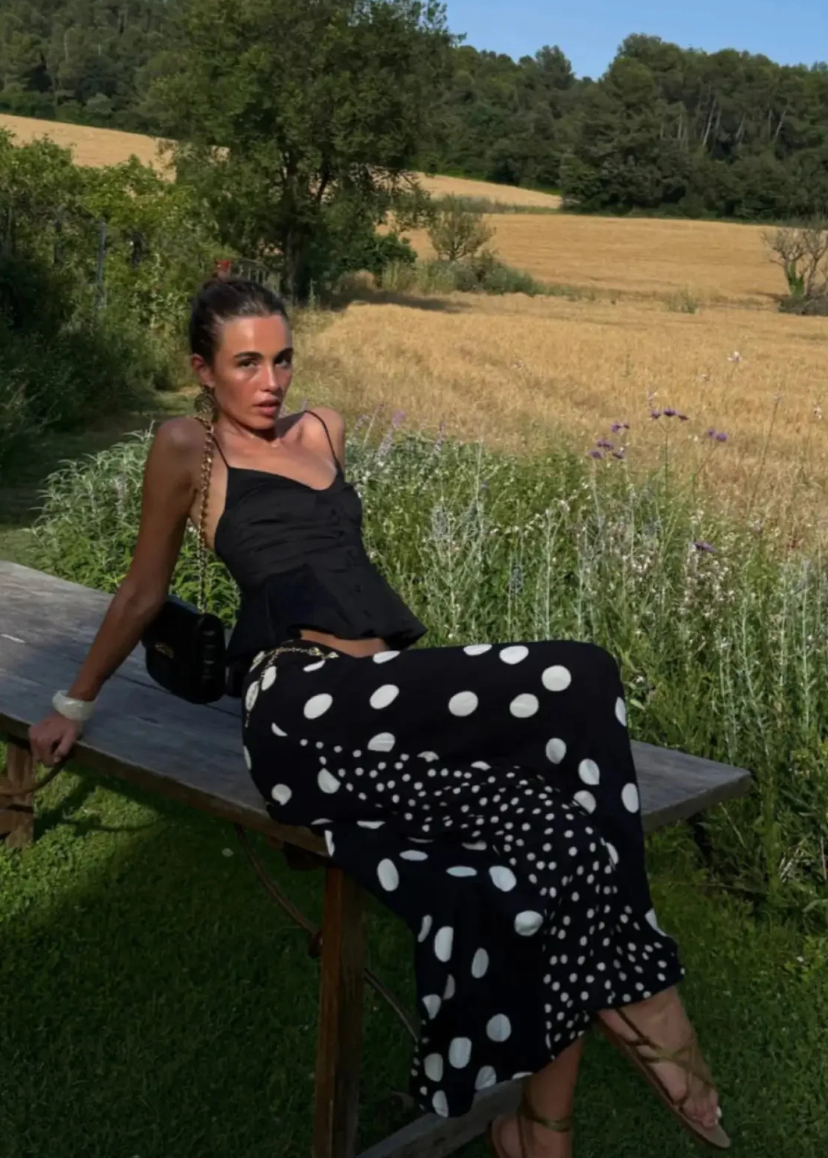 Woman sitting on a wooden bench in a field with a black top and polka dot skirt.