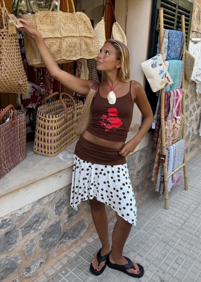 Woman in a brown top and polka dot skirt standing in front of wicker bags and clothing.