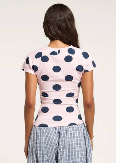 A woman shown from the back wearing a light pink short-sleeve top with large navy blue polka dots, paired with blue and white plaid pants, against a plain off-white background.