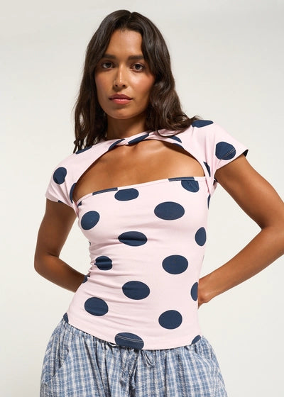 A woman modeling a light pink short-sleeve top with large navy blue polka dots and a square neckline with a cutout detail, paired with a blue and white plaid skirt against a plain light background.