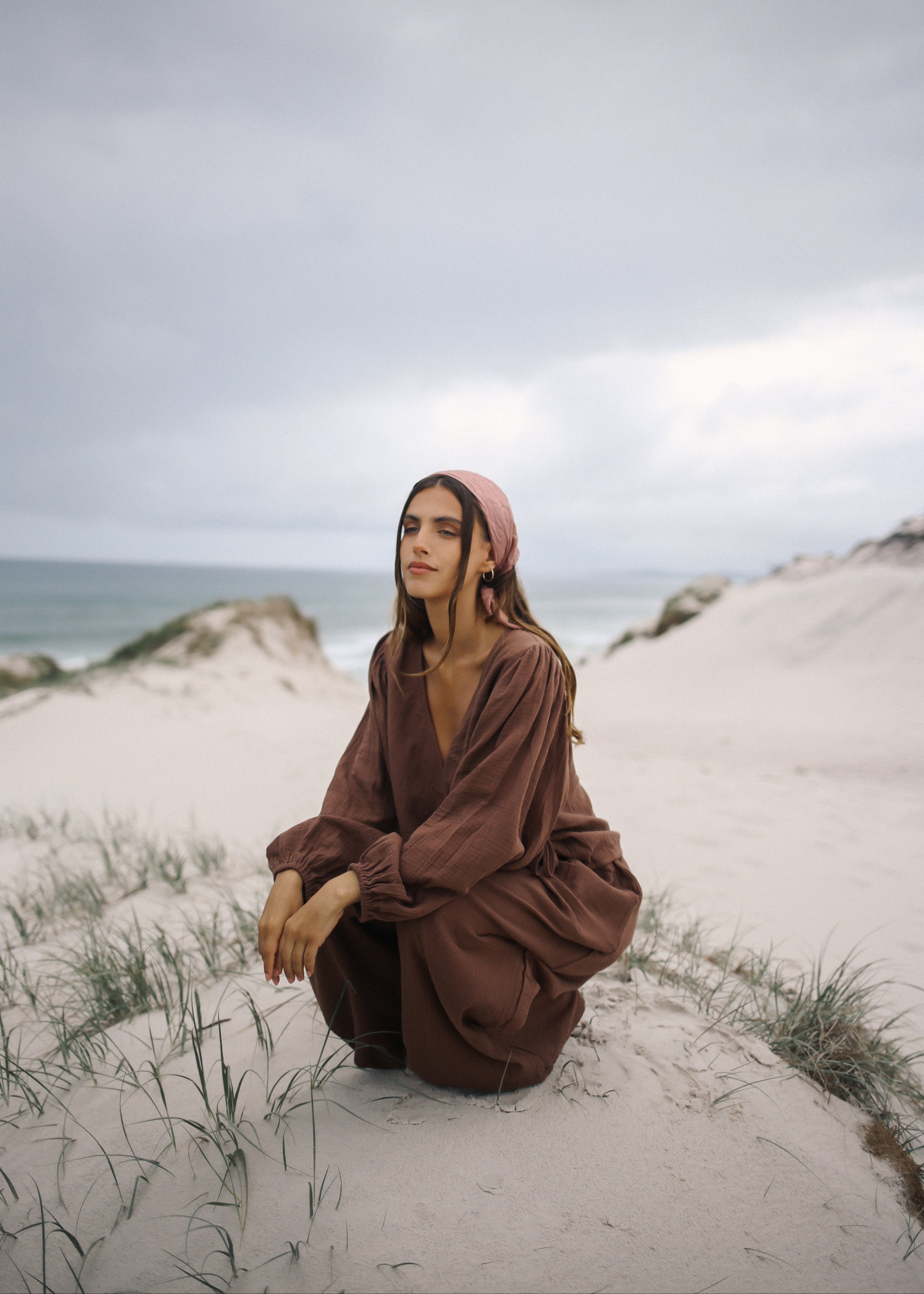 Woman in a brown outfit sitting on a sandy dune with ocean in the background