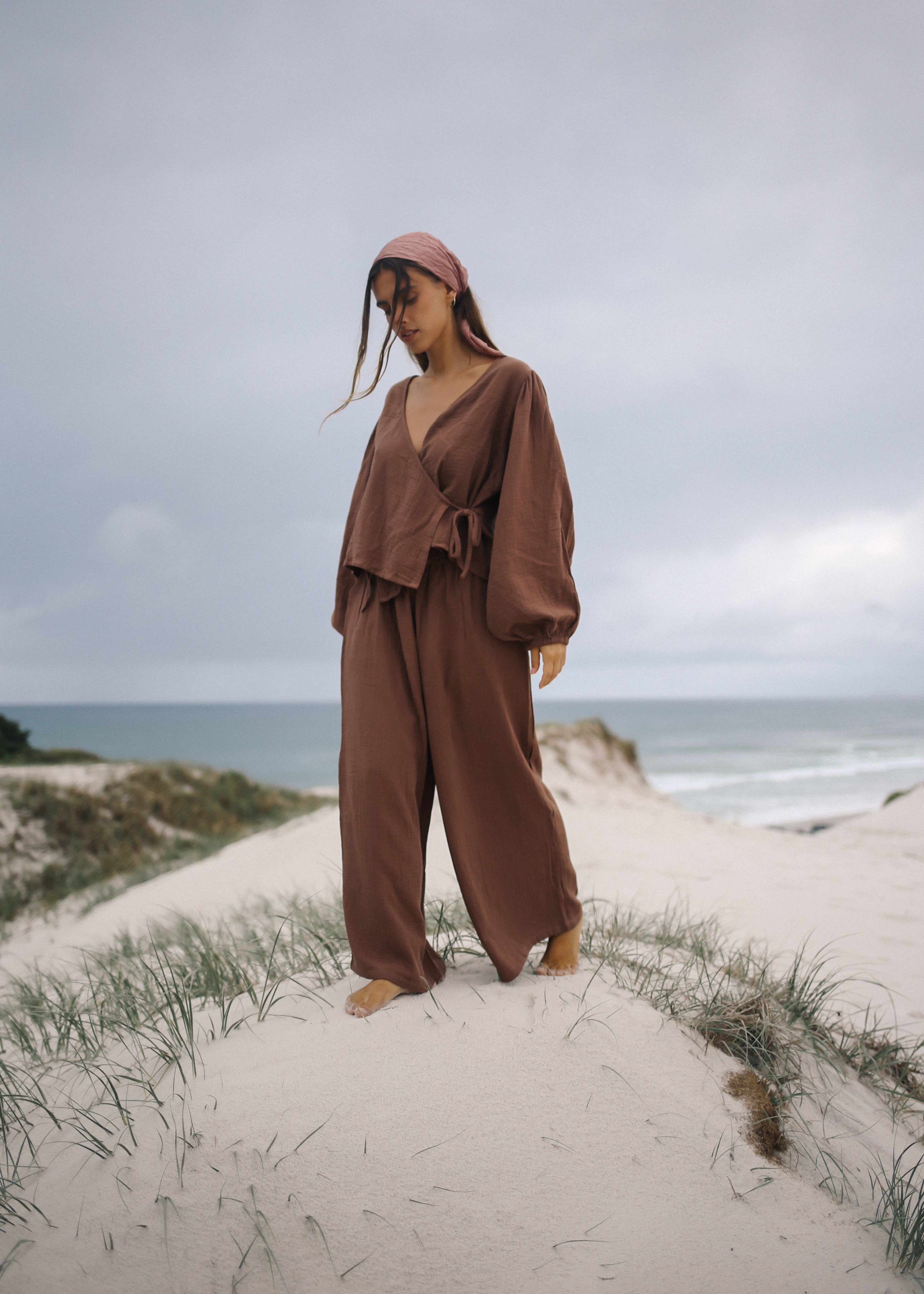 Person wearing a brown outfit standing on a sandy dune with ocean in the background