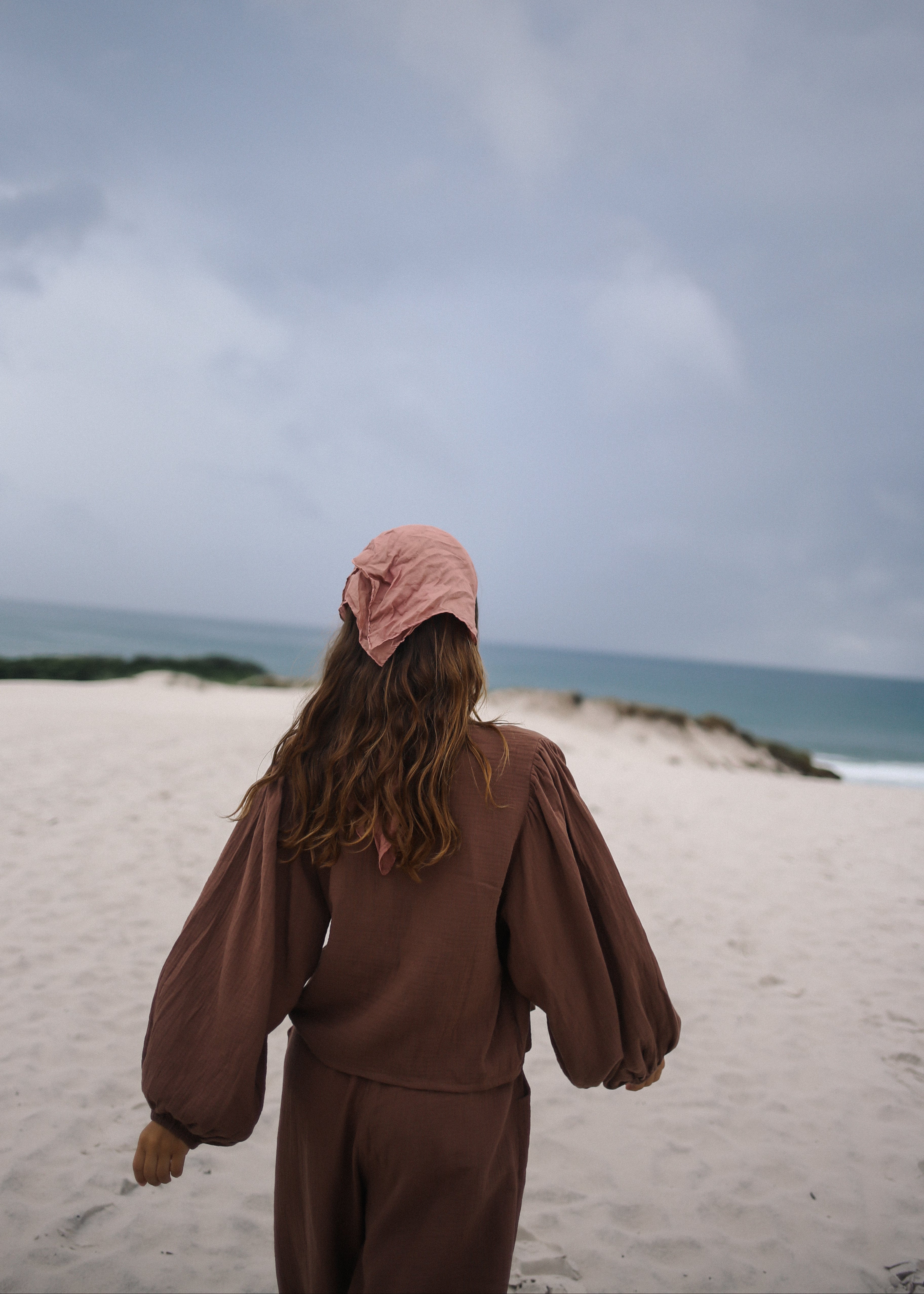 Person wearing a brown outfit and pink hat walking on a sandy beach with ocean and sky in the background