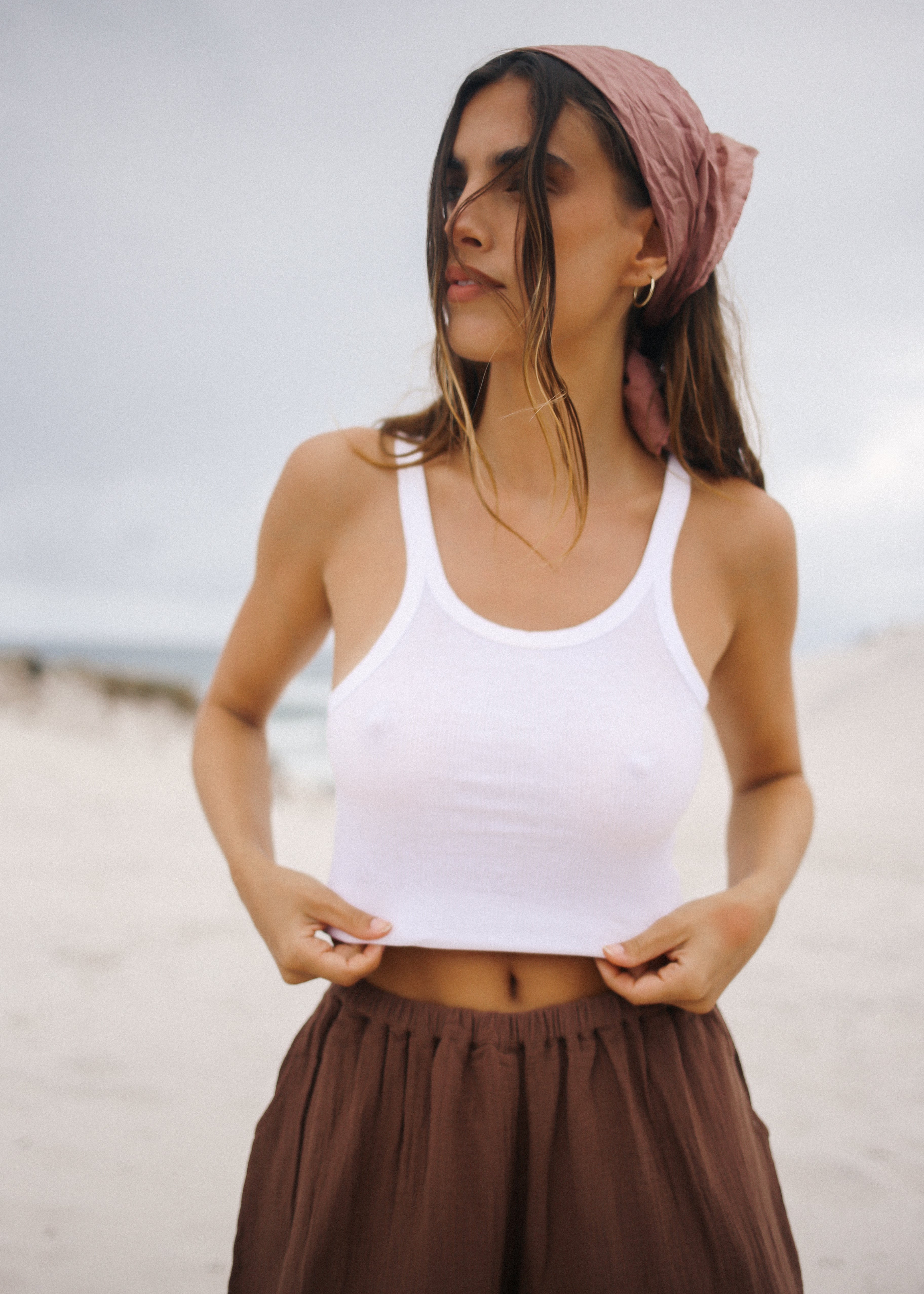 Woman wearing a white tank top and brown pants with a pink headscarf on a beach.