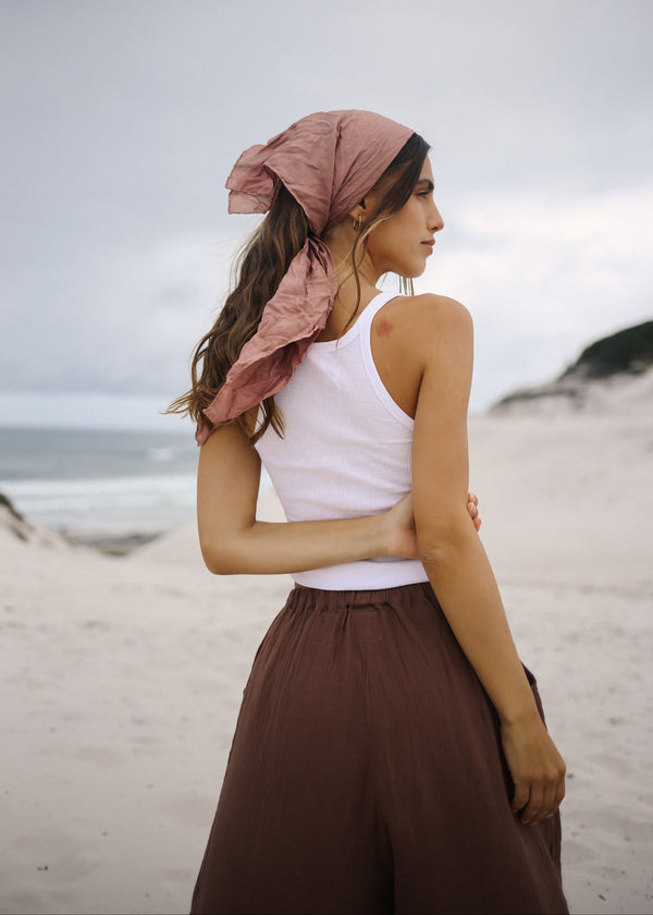 Woman standing on a beach wearing a pink headscarf and white top with brown pants.