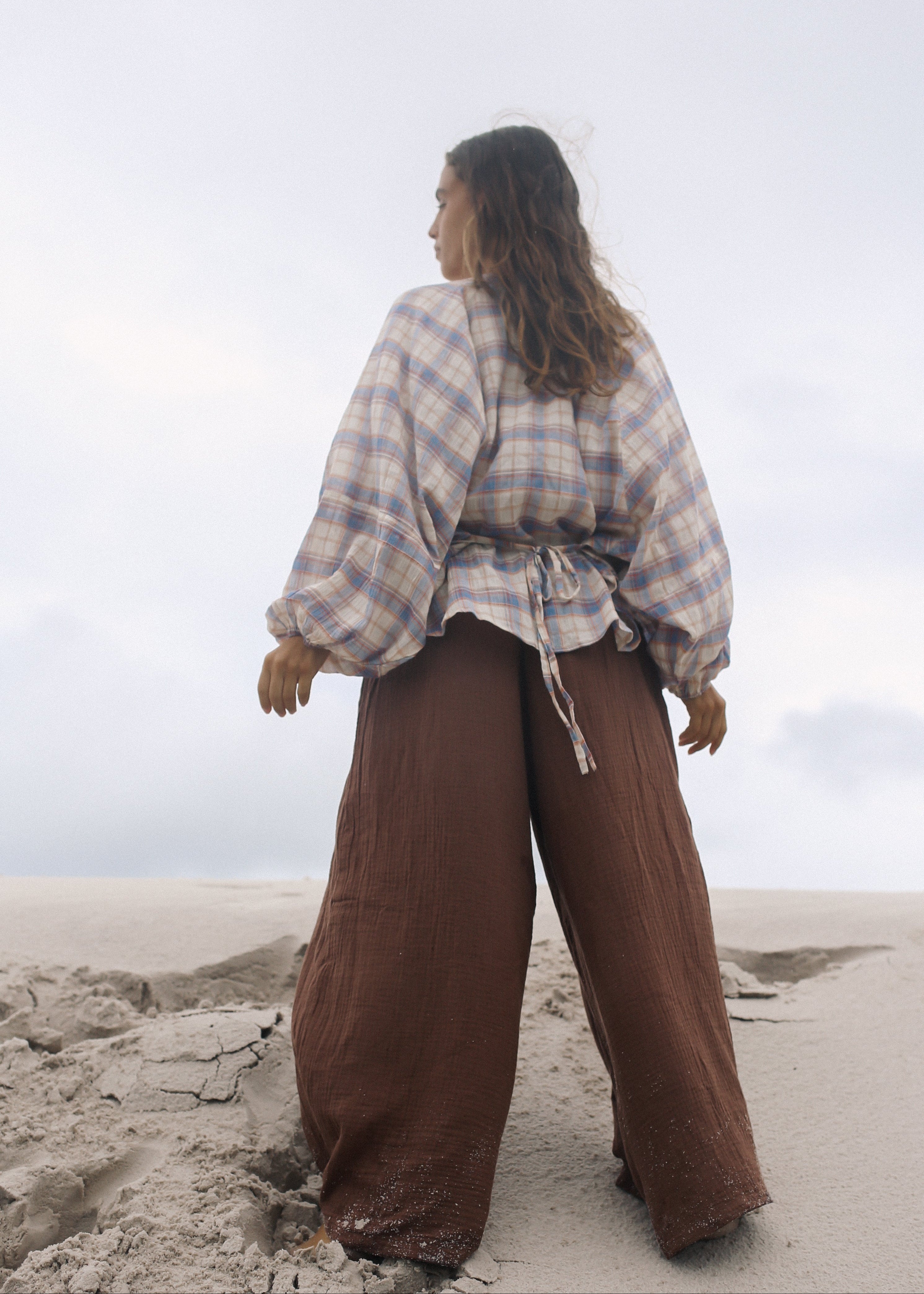 Person wearing a plaid shirt and brown pants standing on a sandy surface with a cloudy sky.
