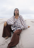 Person sitting on a sandy beach wearing a plaid shirt and brown pants.