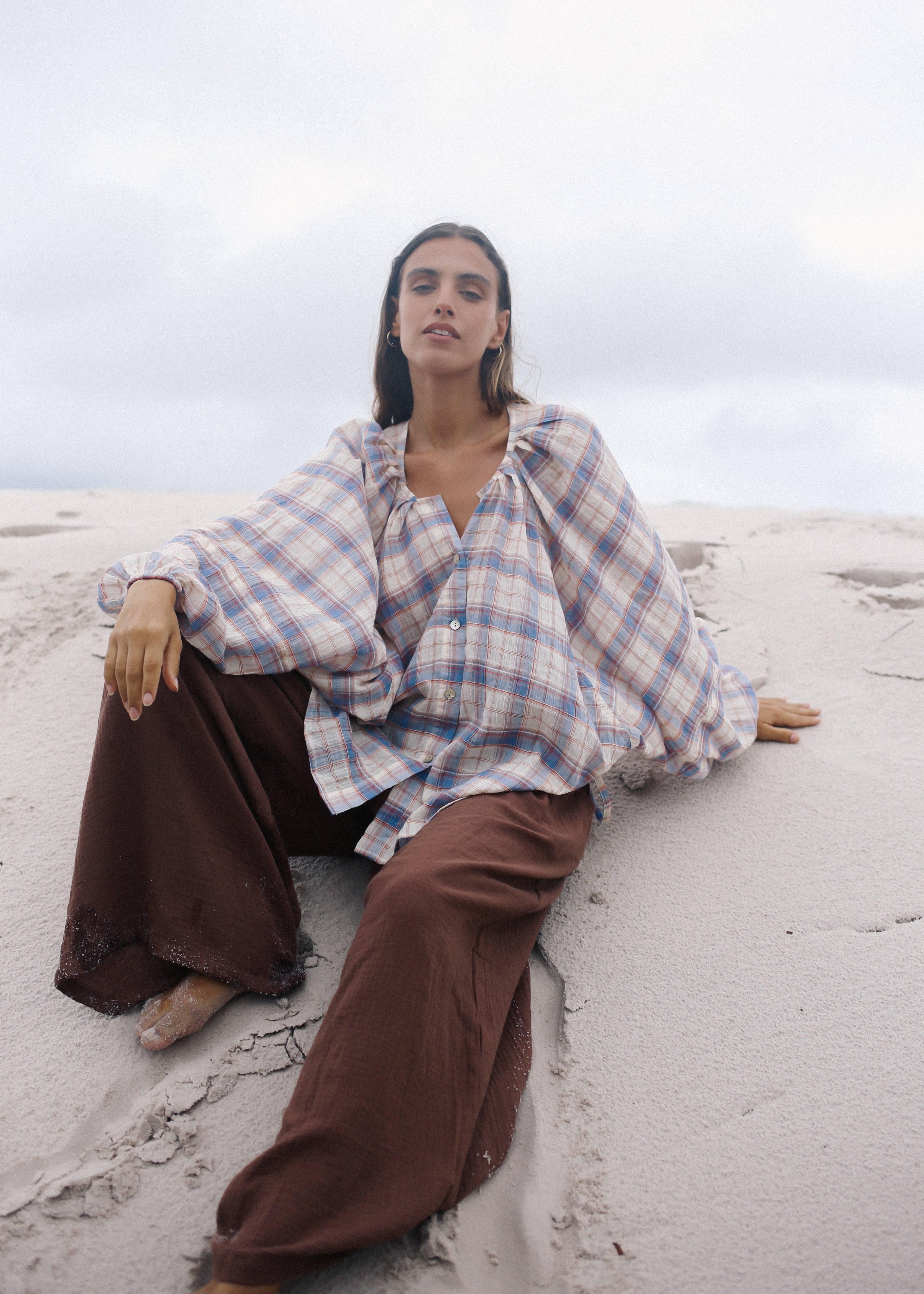 Person sitting on a sandy beach wearing a plaid shirt and brown pants.