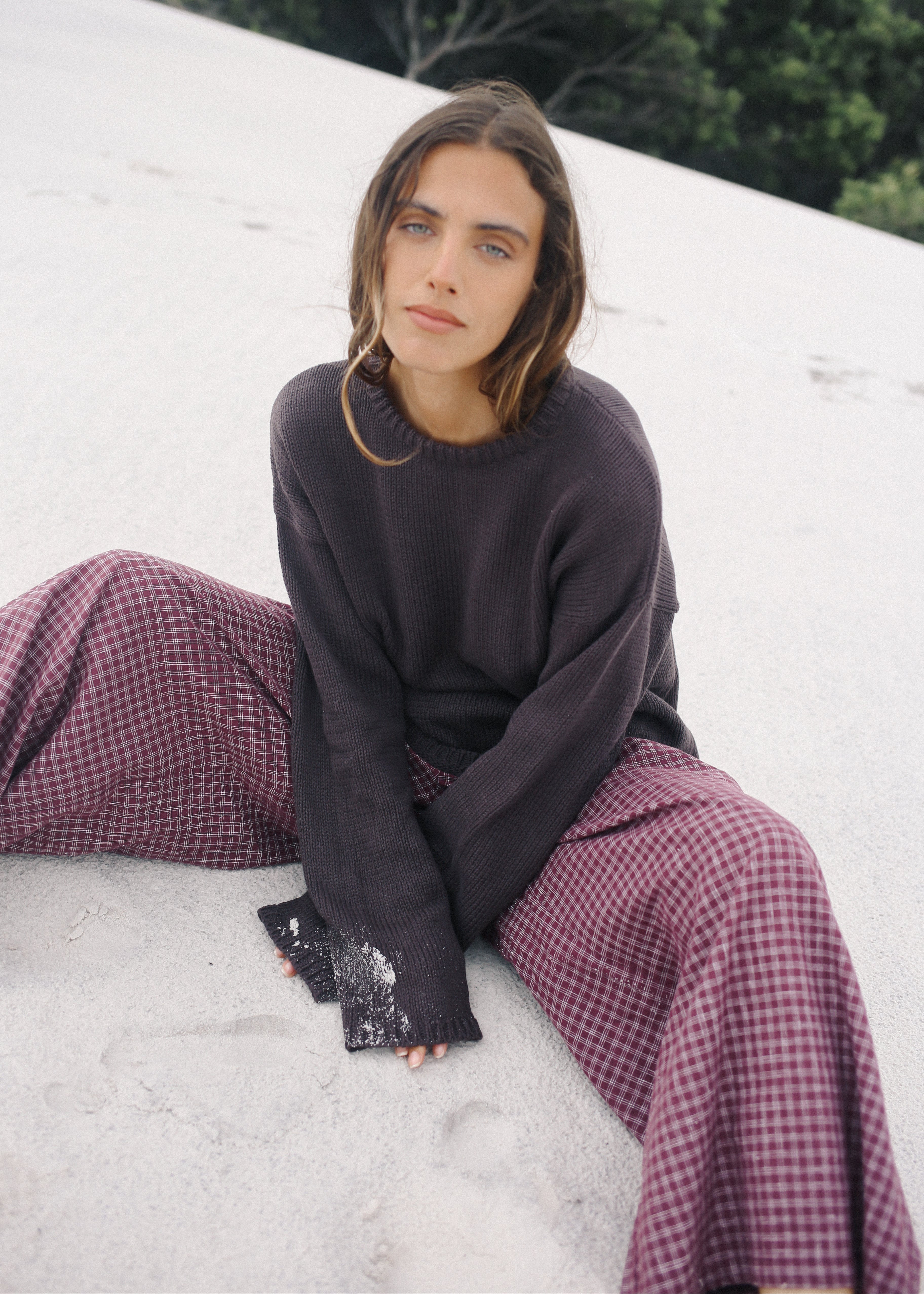 Woman sitting on a white surface wearing a dark sweater and purple checkered pants.