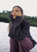 Woman wearing a dark sweater and plaid skirt sitting on a white surface with greenery in the background