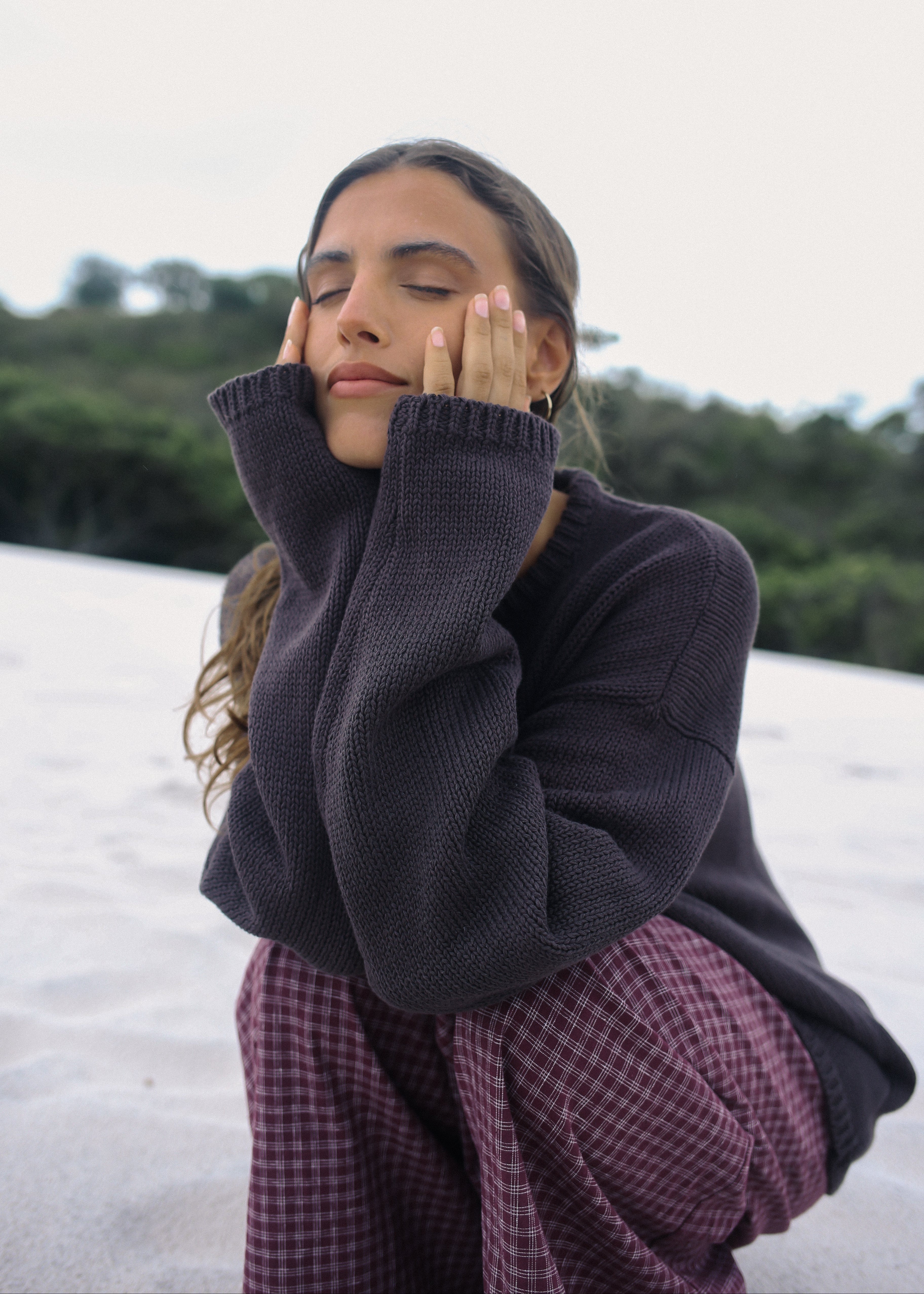 Woman wearing a dark sweater and plaid skirt sitting on a white surface with greenery in the background