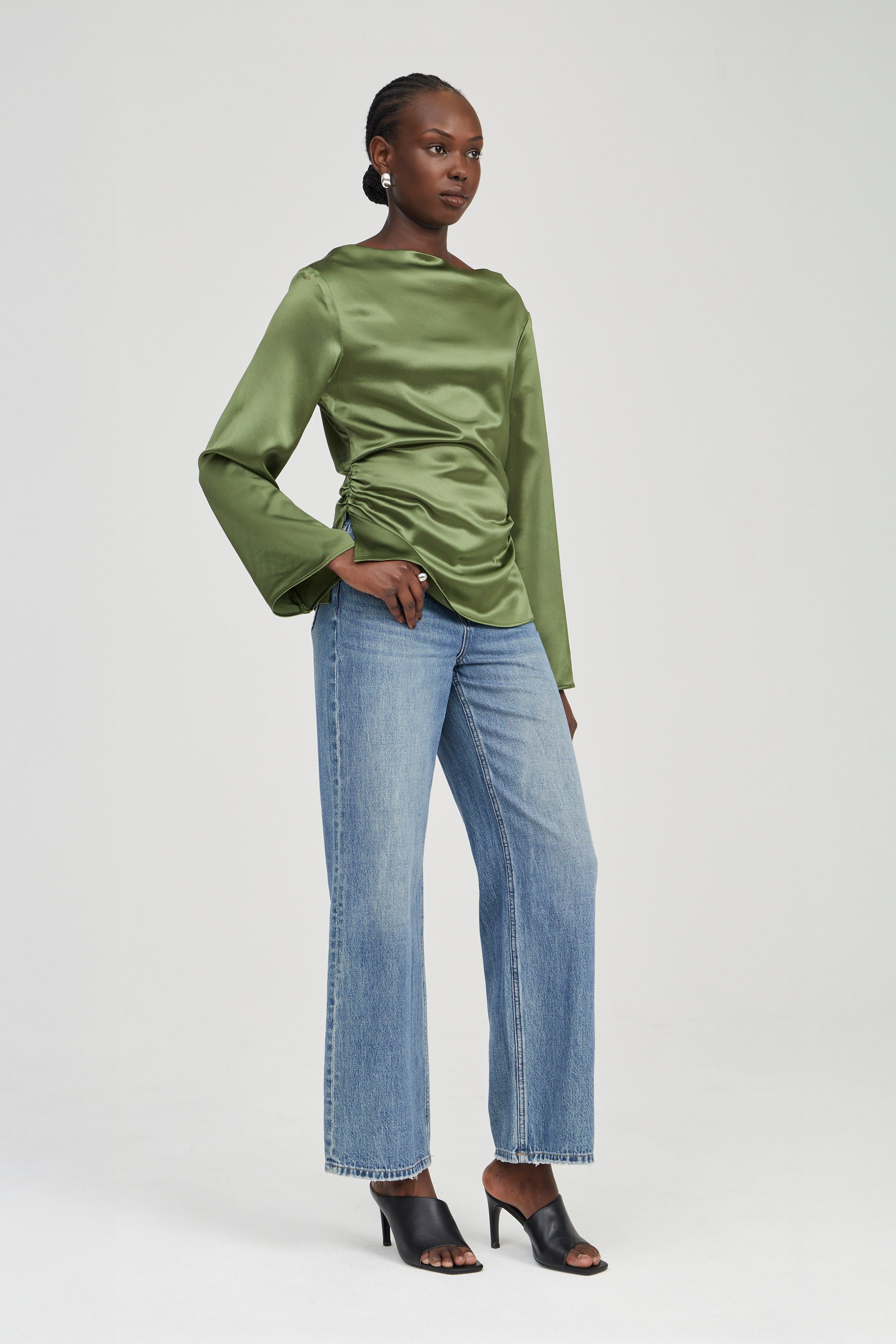 A model wearing a sage green satin long-sleeve top, light blue wide-leg jeans, and black heeled sandals, standing against a light gray studio background.