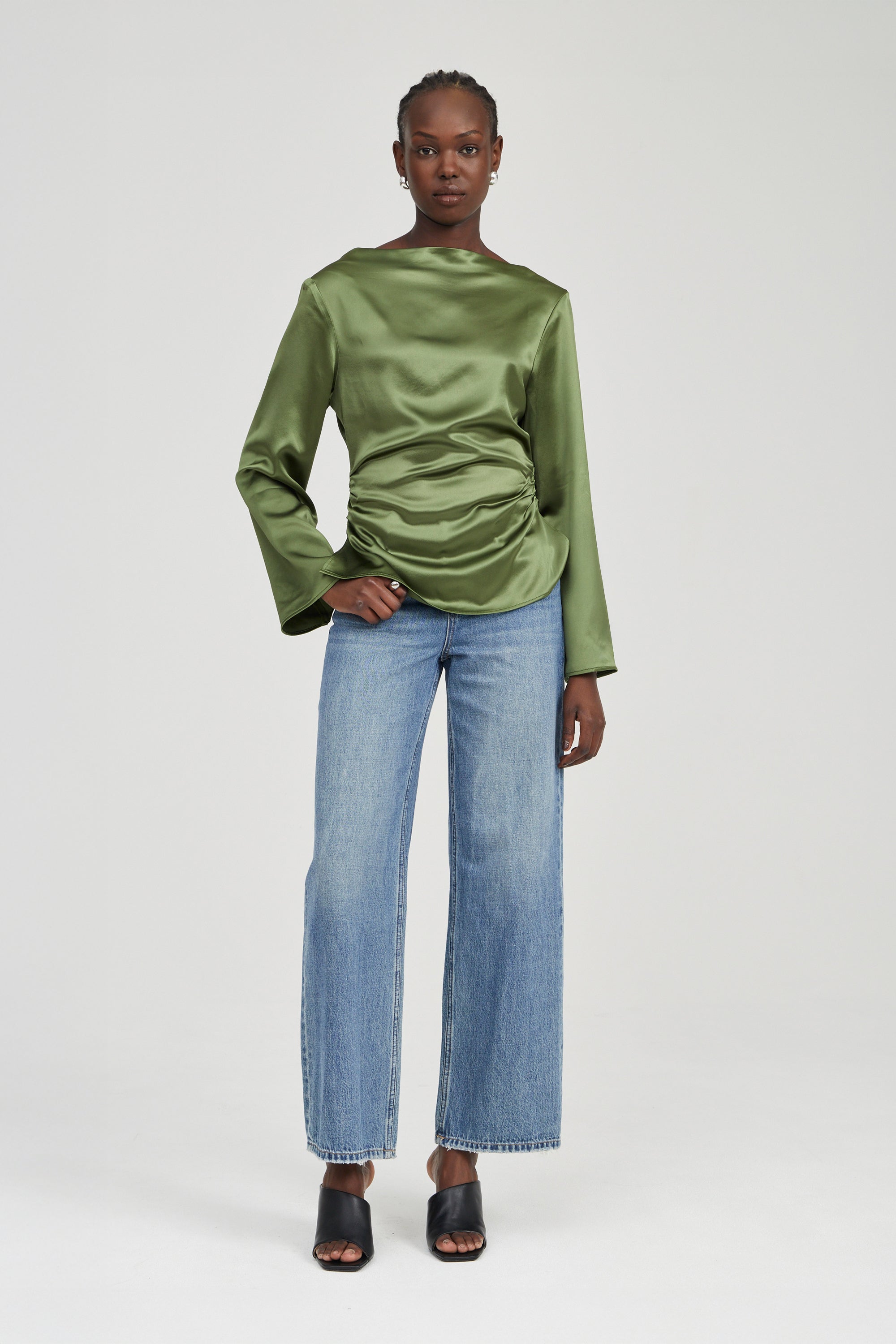 A model stands against a light gray studio background wearing a shiny olive green long-sleeve top with gathered draping at the waist, light blue wide-leg jeans, and black heeled sandals.