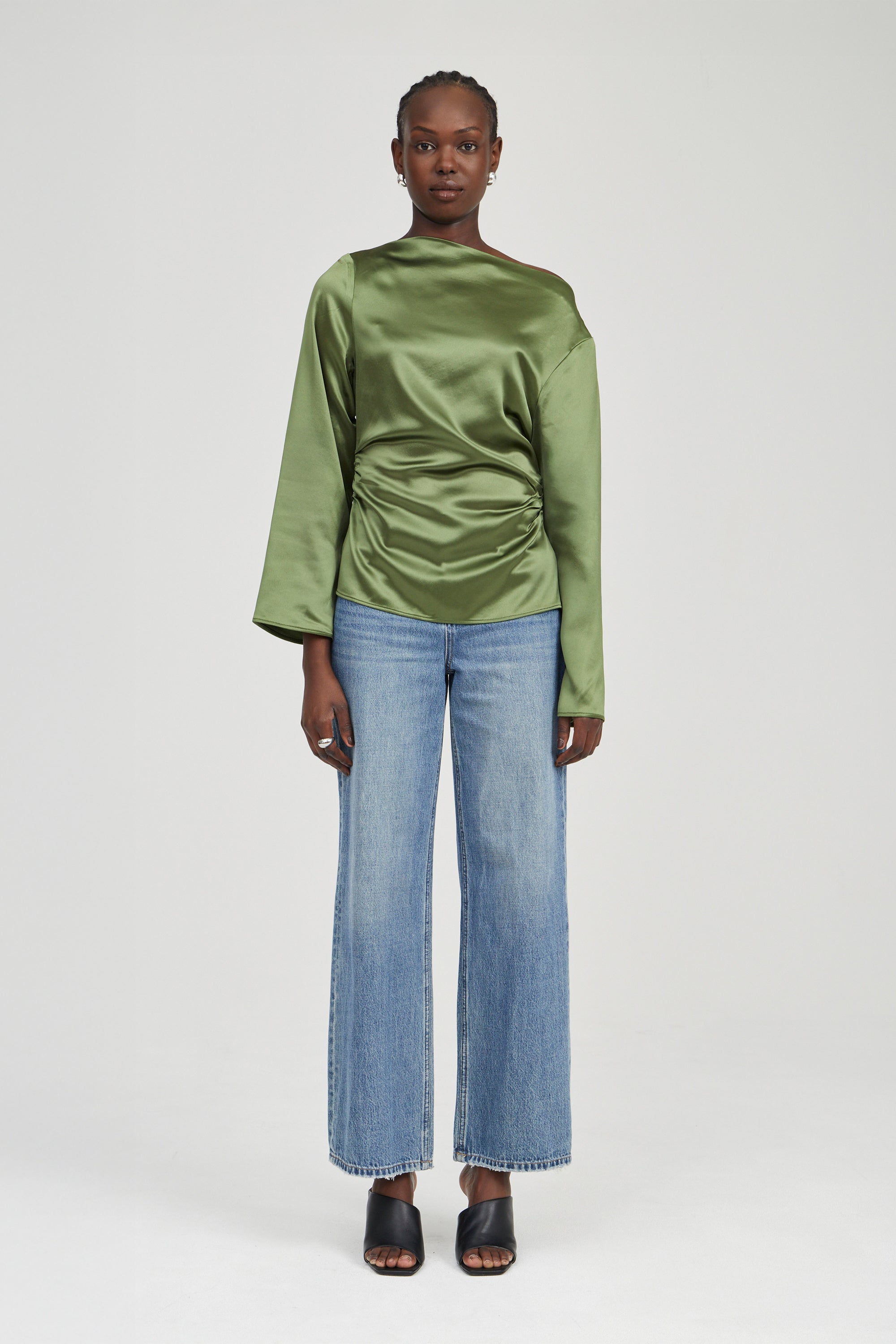A model stands against a light gray studio background wearing a shiny olive green long-sleeve top with gathered draping at the waist, light blue wide-leg jeans, and black heeled sandals.