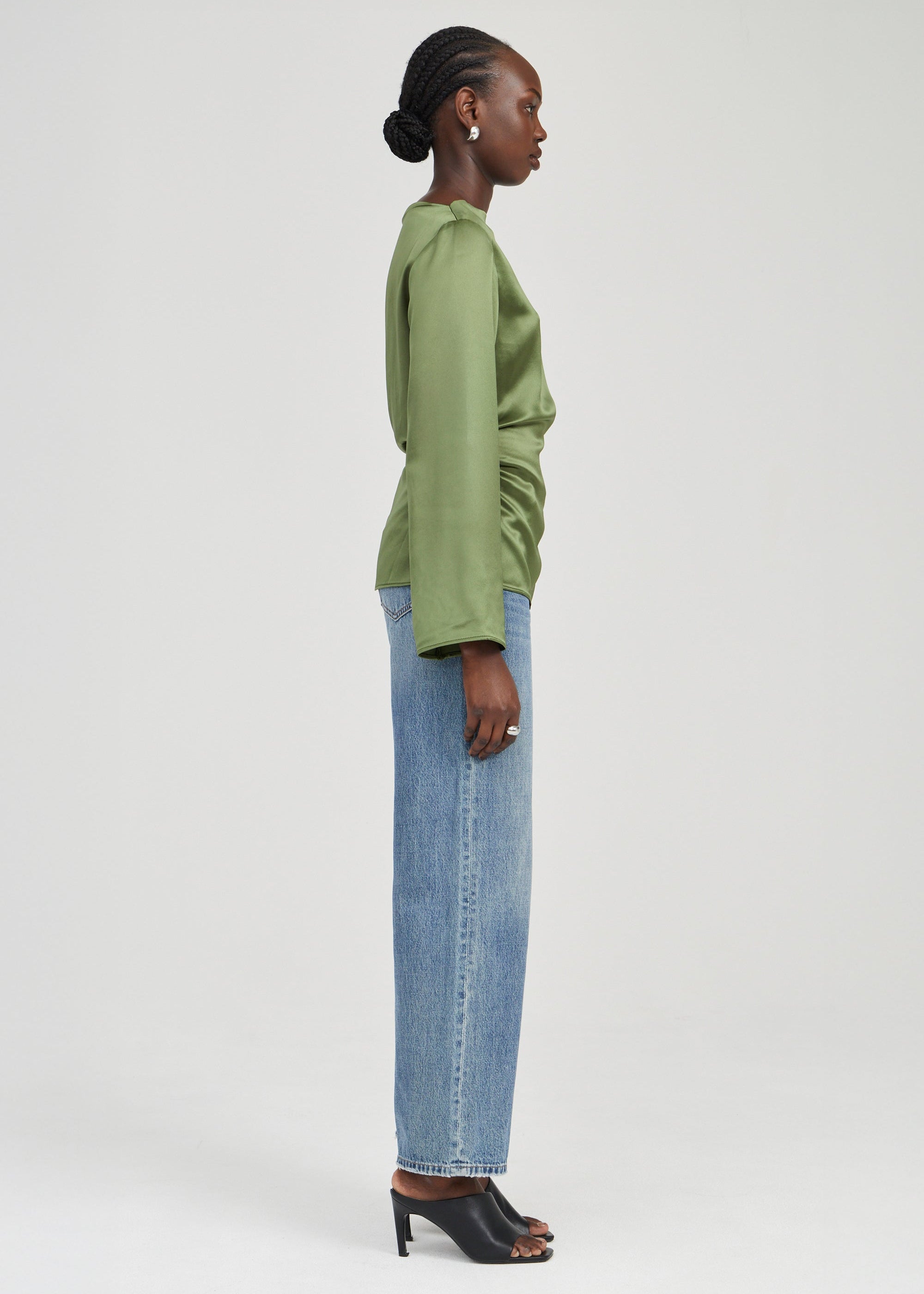 A woman shown in side profile wearing a sage green long-sleeve top, light blue wide-leg jeans, and black heeled sandals against a plain light gray studio background.