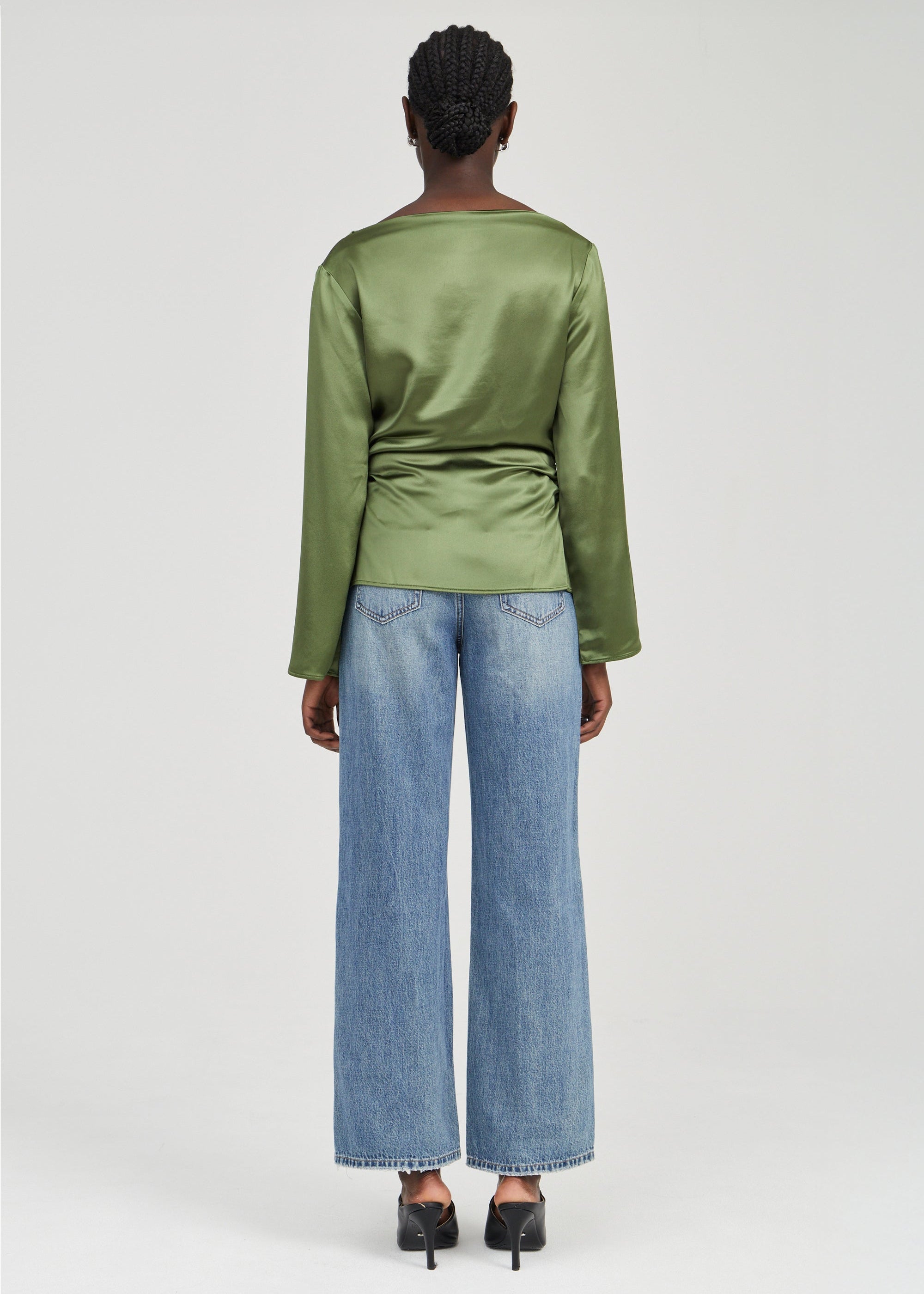A model shown from the back wearing a sage green satin long-sleeve top and wide-leg blue jeans, paired with black heels, against a light gray studio background.