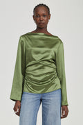A woman modeling a sage green satin long-sleeve top with a gathered ruched front, paired with light blue jeans, against a solid light gray background.