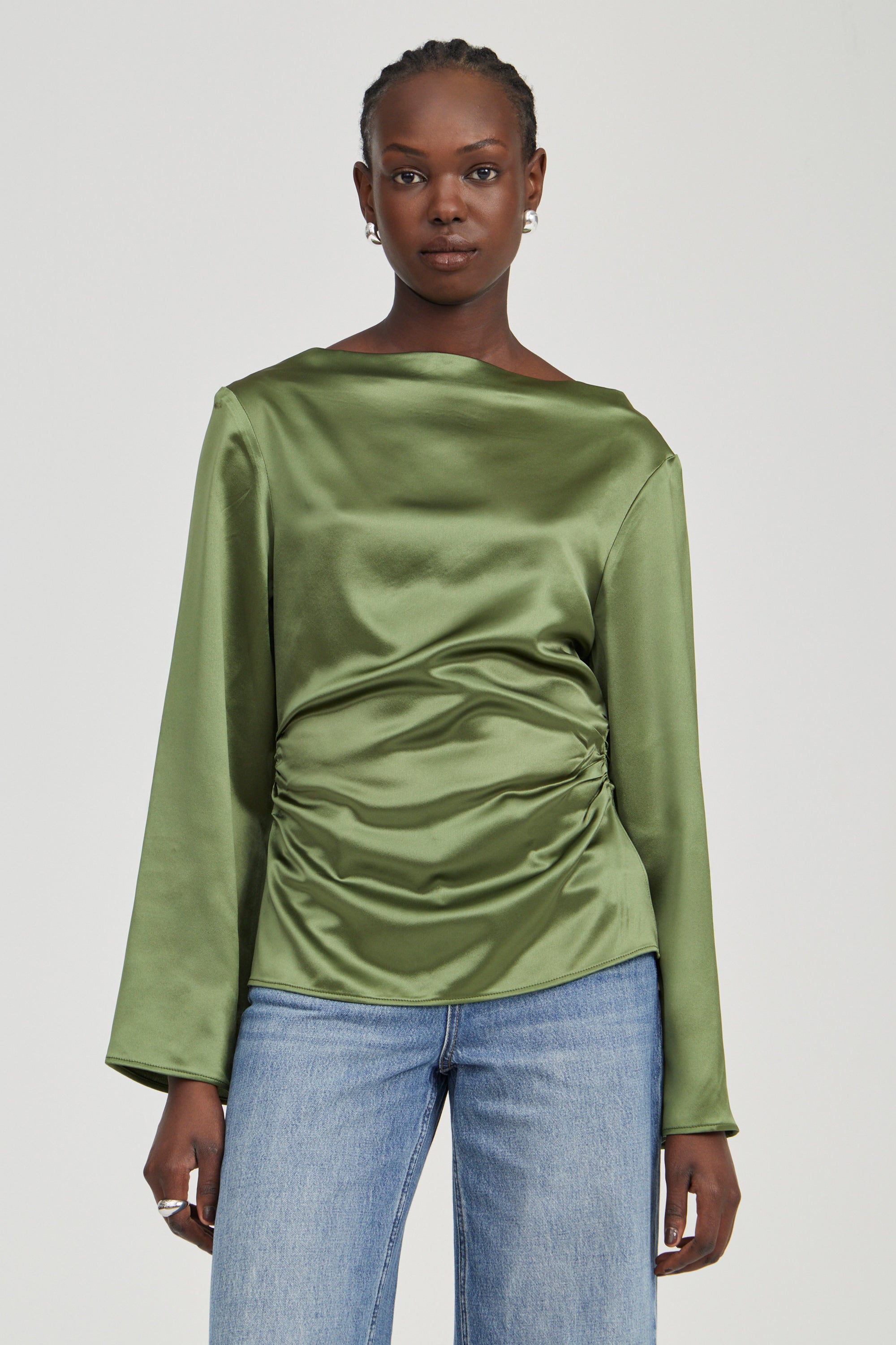 A woman modeling a sage green satin long-sleeve top with a gathered ruched front, paired with light blue jeans, against a solid light gray background.