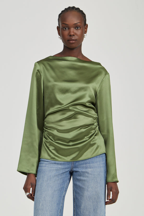 A woman modeling a sage green satin long-sleeve top with a gathered ruched front, paired with light blue jeans, against a solid light gray background.